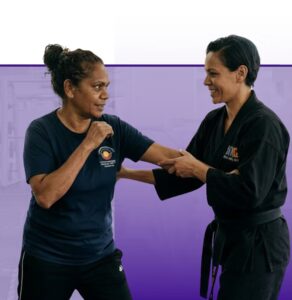 Two women in martial arts attire demonstrate a combat move against a purple wall, representing workplace wellness training