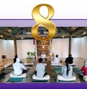 Group yoga class with participants seated on mats in a bright studio with wooden beams and large gold number 8 above