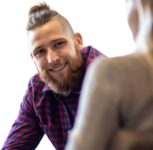 Smiling man with beard and mohawk hairstyle in red and blue plaid shirt, engaged in conversation with another person