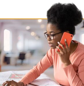 Woman wearing glasses and coral shirt talking on phone while reviewing documents at her desk in a modern office