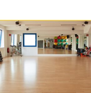 Spacious fitness studio with wooden flooring, wall-mounted mirrors, exercise equipment including stationary bike, colorful stability balls, resistance bands, and training props organized along the walls