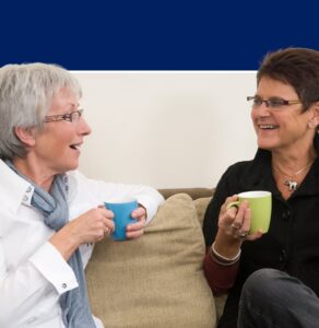 Two women sitting on a couch smiling and holding mugs, representing workplace connection and wellbeing