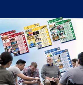 Group of adults discussing colorful program posters displayed outdoors, including DISC personality and workplace wellness training materials