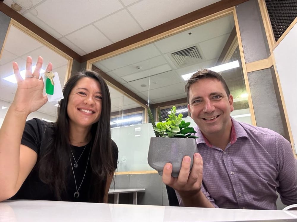 Two smiling team members holding a potted succulent plant in an office, one waving at the camera