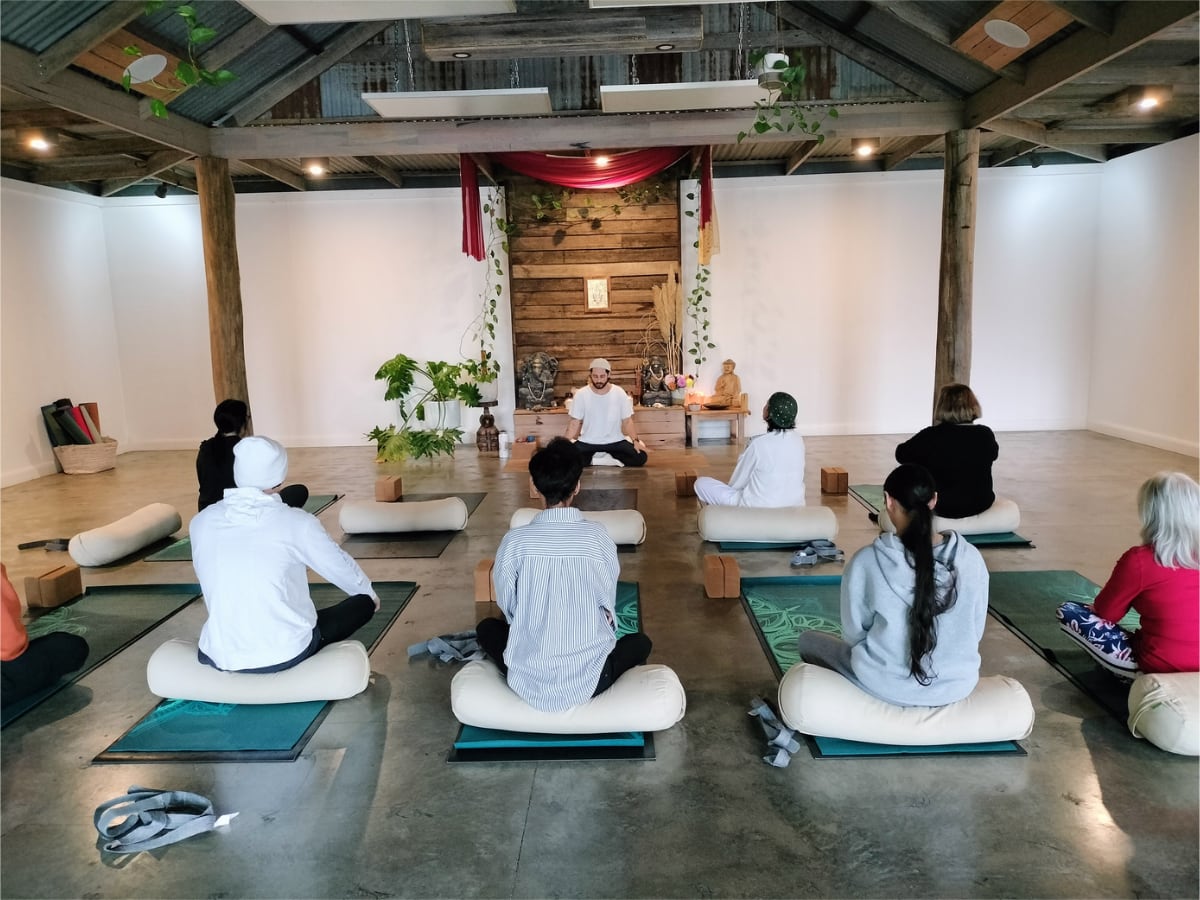 Yoga class in session with instructor leading group of participants seated on mats in a rustic studio space with exposed beams and wooden backdrop